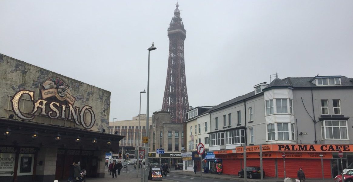 Photograph showing Blackpool Tower against a grey sky. In the foreground is a grey 'Coral Island Casino' on the left of a crossroads; on the right is 'Palma Cafe' closed with the orange shutters down. The photograph could be described as creating an atmostphere that captures a drab, dull and garish scene at the same time. The streets depicted are not quite deserted - a few people seem to be going about their daily business.