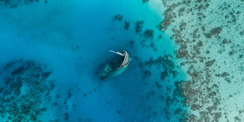 An aerial shot looking down at beautiful clear blue-green waters. In the centre of the photograph is a shipwreck - one end of the ship poking out above the water. You can just make out a person swimming towards it.
