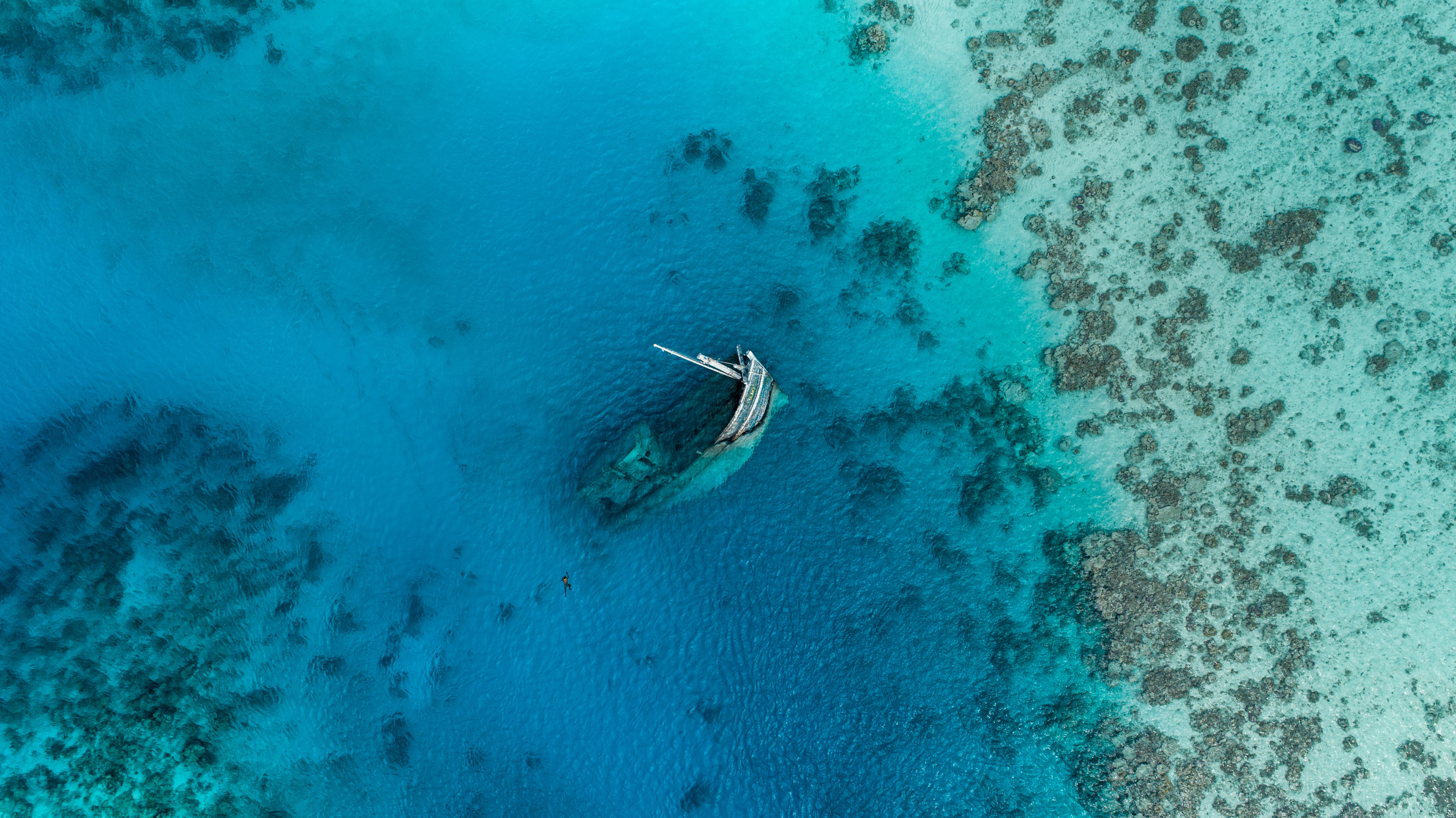 An aerial shot looking down at beautiful clear blue-green waters. In the centre of the photograph is a shipwreck - one end of the ship poking out above the water. You can just make out a person swimming towards it.